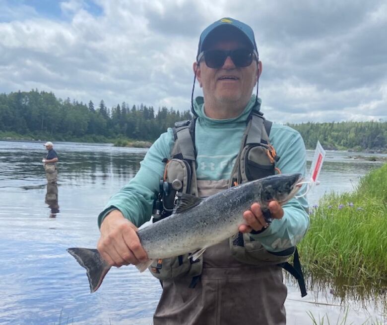 A man wearing hip waders stands smiling in a river holding a tagged salmon. 