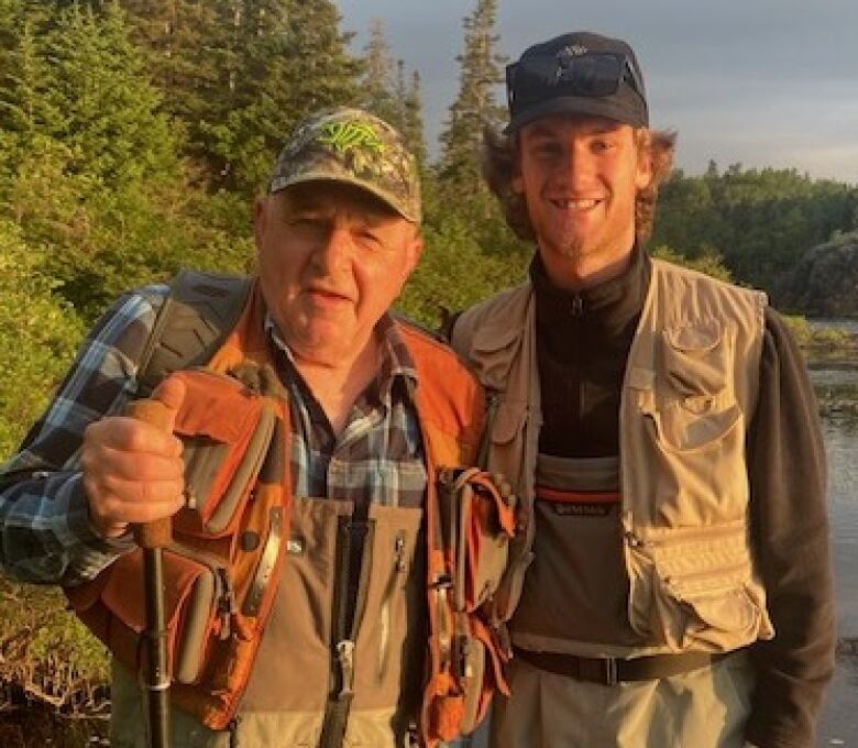 Two men wearing fishing gear smile while standing next to a river.