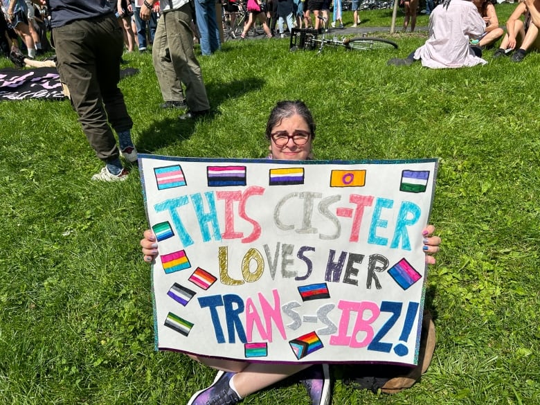 A woman sits cross-legged in the grass holding a sign that reads, 