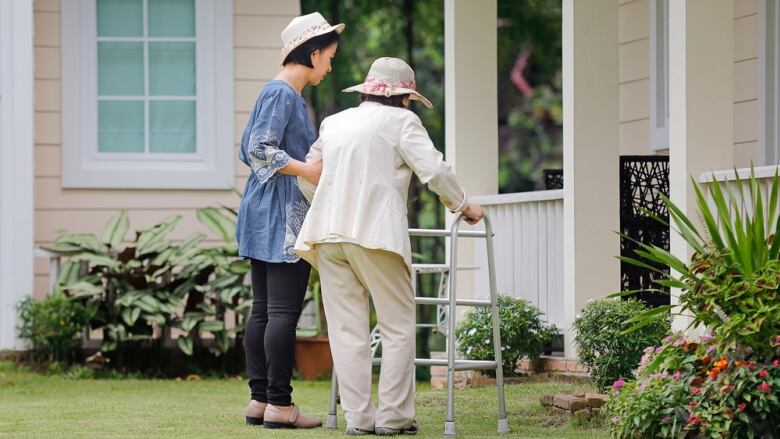 A younger woman helps an older woman with a walker walk in a yard outside a home.