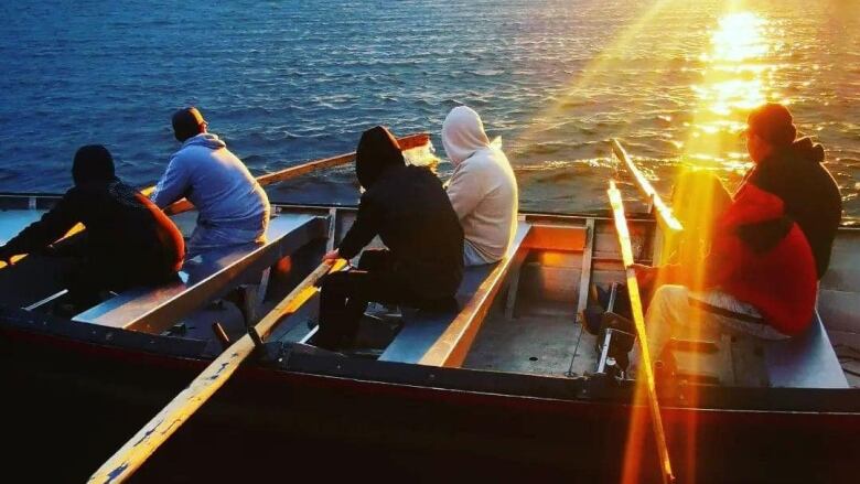 Six rowers in a large boat watch the sunset. 