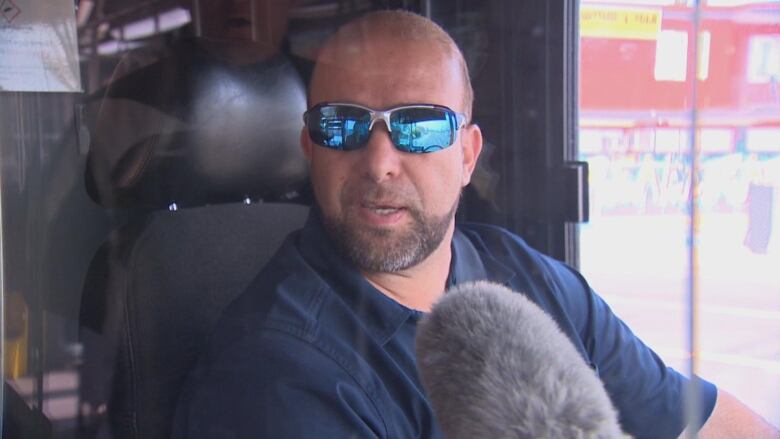 A bus driver wearing sunglasses sits in the drivers seat of a bus.