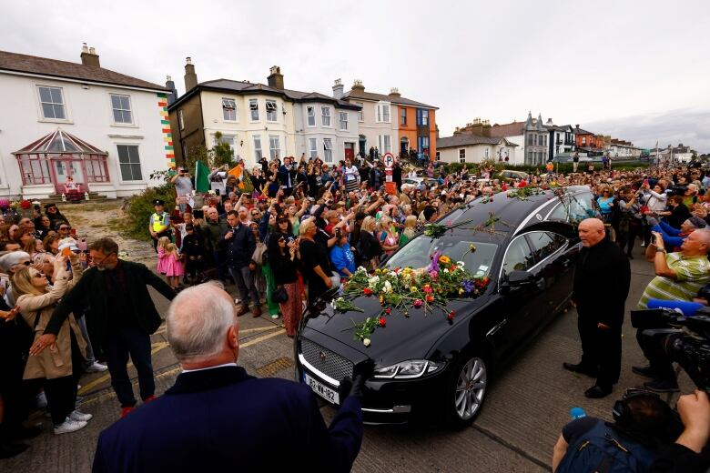 A crowd of people surround a hearse during a funeral procession.