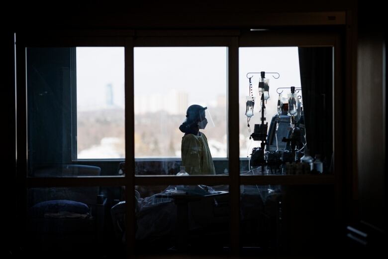 Nurse in safety gear stands next to hospital bed and IV drips, backlit against window