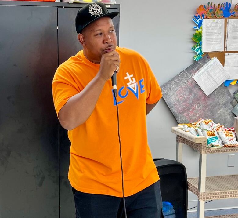 A man wearing an orange shirt and ball cap speaks into a microphone.