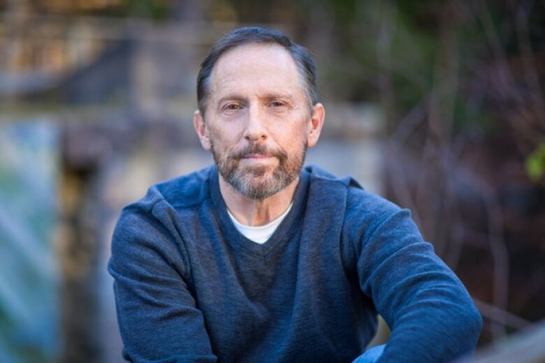 A man with a beard, greying hair and a serious expression on his face. He's wearing a blue, v-neck sweater and the background is blurred. 