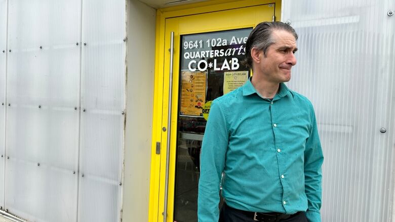 A man in a blue collared shirt stands before a bright yellow door. The building behind him is a metallic silver.