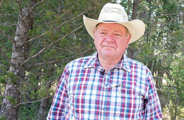 A man surrounded by trees in a white cowboy hat and a blue-and-white checked button-up shirt with an open collar looks at the camera.