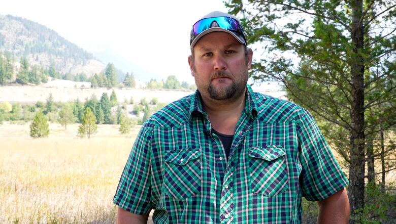 A bearded man in a dark green striped button-up shirt wearing a baseball cap with sunglasses resting on the brim stands in a field.