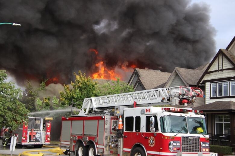 Flames erupt from behind a series of roofs, with a fire truck in the foreground.