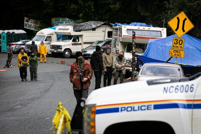 Camper vans and RCMP SUVs are seen on a road in a forest.