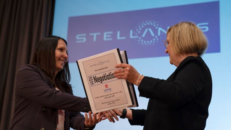 Two woman shake hands while holding a binder