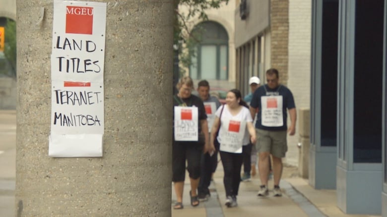 A sign posted on a concrete column says Land Titles/Teranet Manitoba. A group of people are walking a picket line in the background.