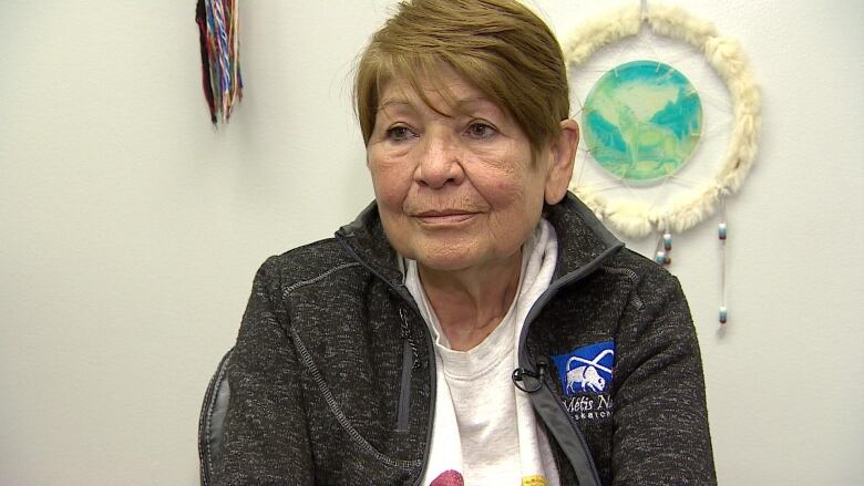 A woman can be seen wearing a black jacket over a white T-shirt. There is a dreamcatcher on the wall behind her. 