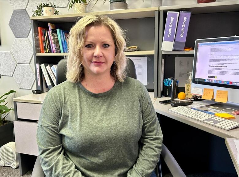 A woman can be seen wearing a green top, she has blond hair and is sitting in front of a desk in an office.