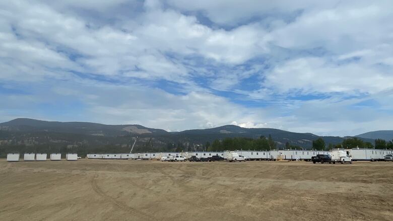 A line of modular housing under a cloud-filled blue sky is seen in the distance across a field.