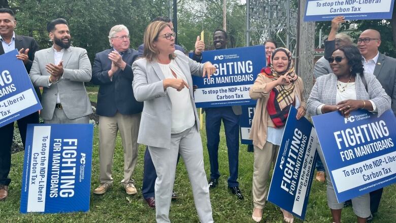 A woman in a suit making hand gestures near other people carrying signs reading 