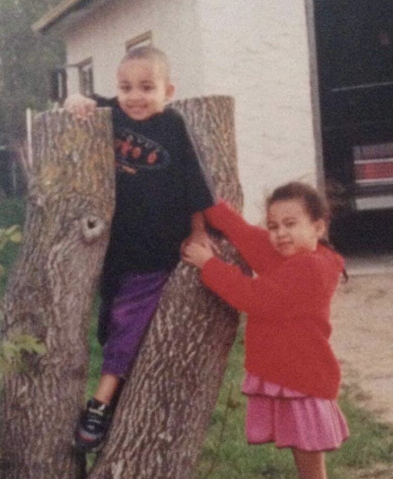 A young boy stands between two tree trunks next to a young girl, who stands beside him.