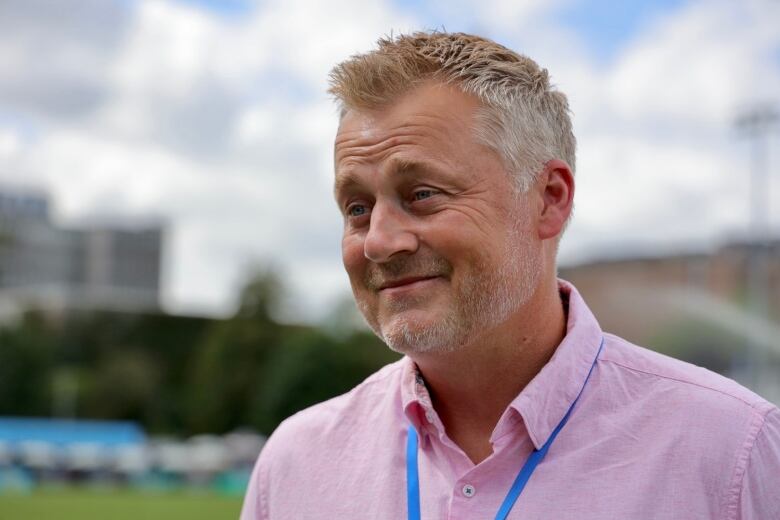 A man wears a light pink shirt and a blue land yard around his neck. He has light blonde and grey hair. 