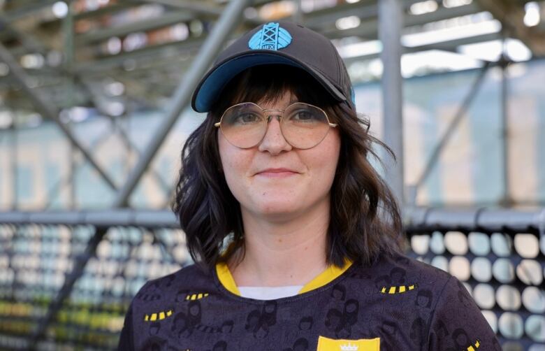 A woman with round gold classes smiles at the camera. She is wearing a black and blue ball cap and a black and yellow soccer jersey. 