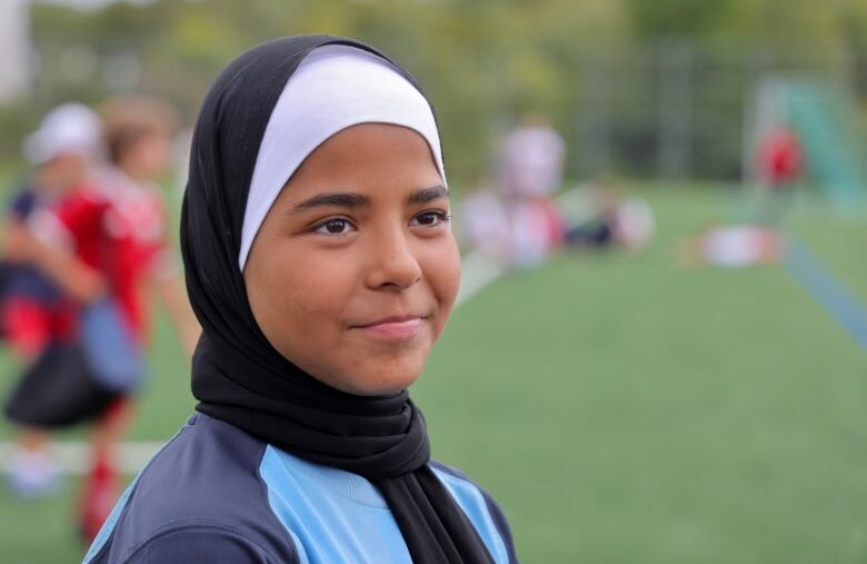 A young girl smiles. She wears a black hijab and light blue soccer jersey. 