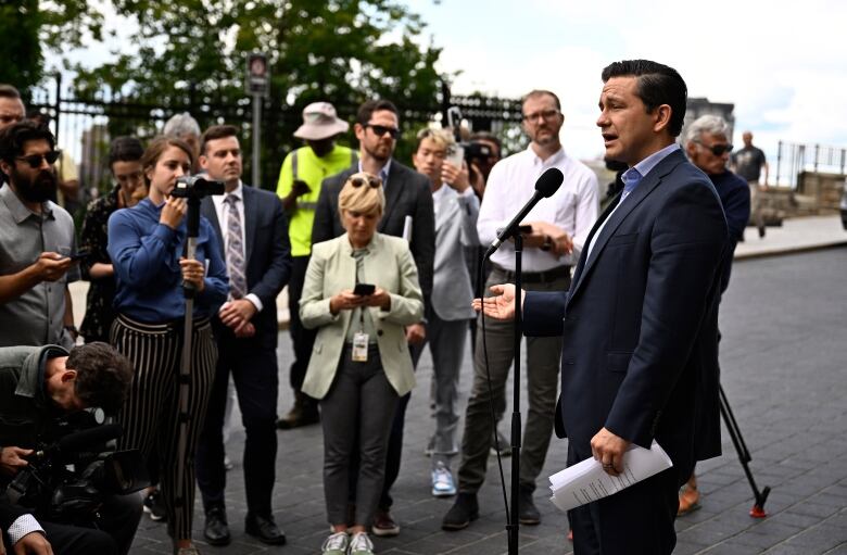 A man in a suit gestures as he speaks in front of a microphone set up on a paved area outside, as a group of reporters stand nearby, taking notes.