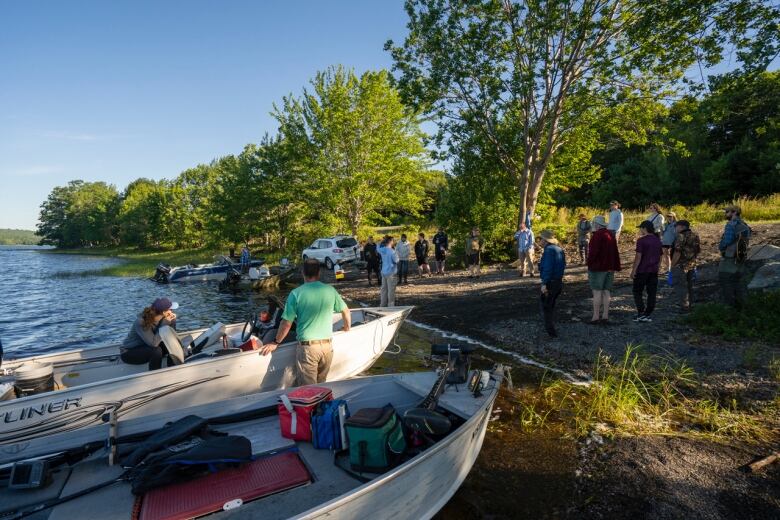 Some people sit in boats on a river bank while others stand nearby.