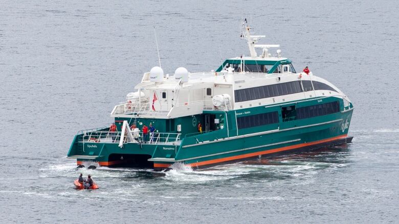 A ferry sits in the open ocean. It is green and white and a small orange zodiak is buzzing behind it with two men in lifejackets.