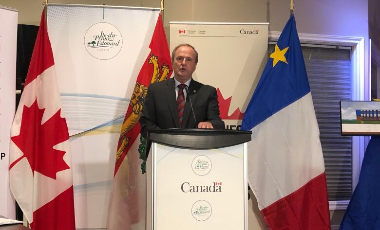 Man in suit stands at podium with Canadian, P.E.I. and Acadian flags around him.