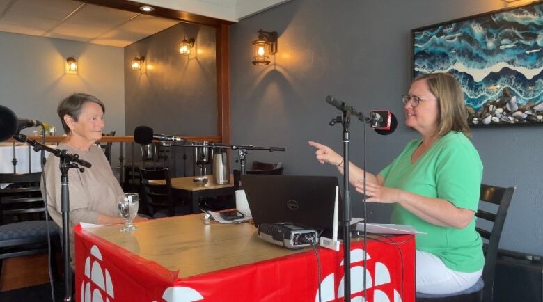 A woman with short brown hair speaks with another woman sitting across the table from her. The table is covered with pieces of radio gear to air a live radio show.