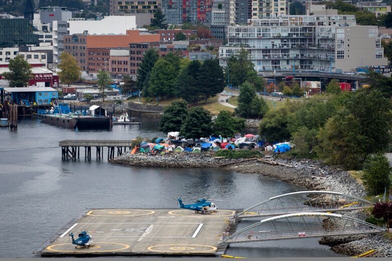 Wide view of helicopters parked in a helipad, and colorful tents at CRAB Park with Downtown Vancouver in the background. 