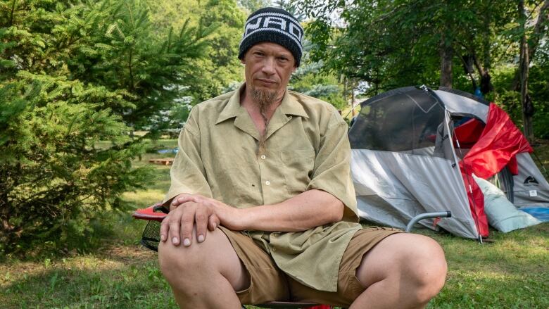 A man sits on a camping chair outside. Behind him is a grey and red tent.