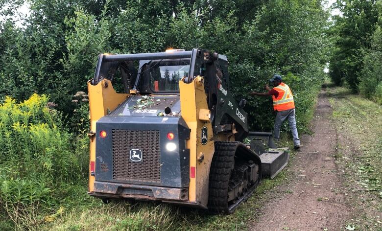 A John Deer small bulldozer, seen from the back. It is sat along the grassy edge of a trail. A person in an orange neon safety vest is positioned ahead of the vehicle.