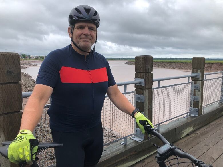 A man wearing a bicycle helmet and standing on a bridge. He is holding the handle and seat of a black bike with his neon gloved hands.