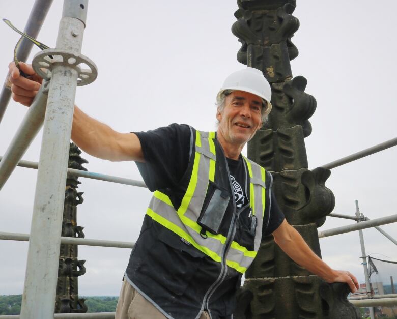 An older gentleman in a hard hat and black t-shirt stands on the pinnacle of a church. 