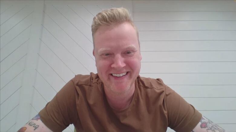 A man in a brown tshirt with blonde hair sits, smiling at the camera. 