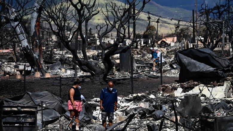 A woman and a man wearing face masks stand in the burned rubble of a home with charred trees behind them. 