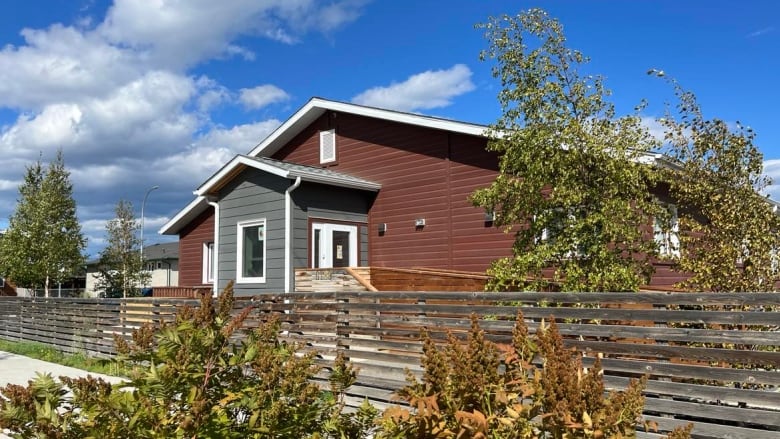 A small building sits behind a wooden fence and some bushes and trees.