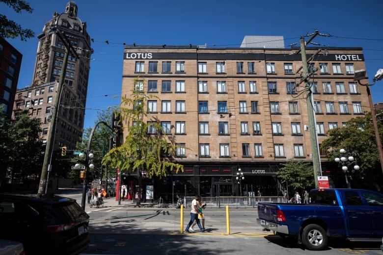 A five-storey building with a brownstone facade has the words Lotus and Hotel at the top of the building in opposite corners. A truck drives by while people walk past with a highrise visible to the left.