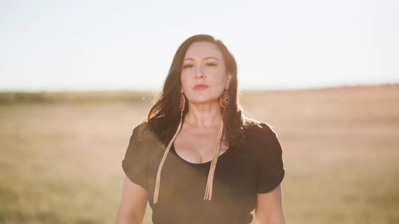 Leela stands in a sun-drenched field wearing a black top and long beaded earrings.