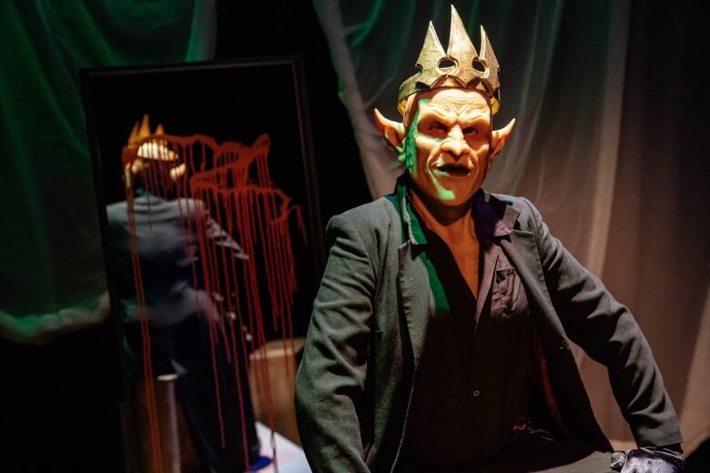 Half-body photo of a performer on stage wearing a realistic goblin mask and crown. A mirror behind them is splattered with blood.