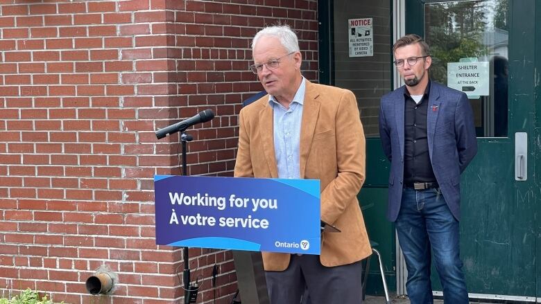 A man stands at a microphone outside. Another man stands behind him.