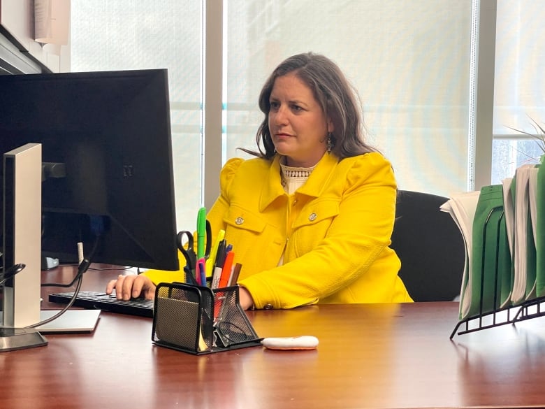 A Caucasian woman in a yellow blazer sits at a desk working on a computer.