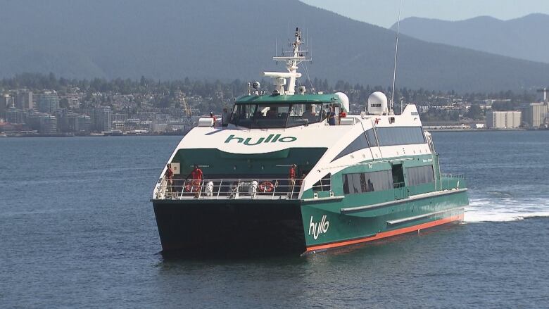 A green and white ferry is pictured pulling up to a dock with city buildings behind it.