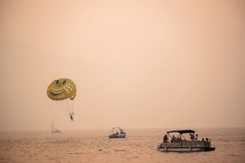 A parasailer with a yellow happy face parachute and three small pleasure boats are shown on a lake. The sky and water are tinted a pinkish orange from wildfire smoke. 