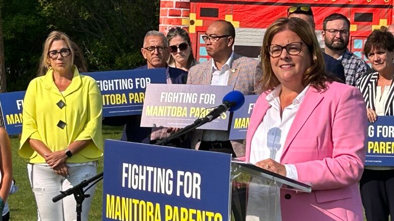 A woman is standing at a lectern speaking into a microphone. Behind her is a crowd of people holding signs that say 