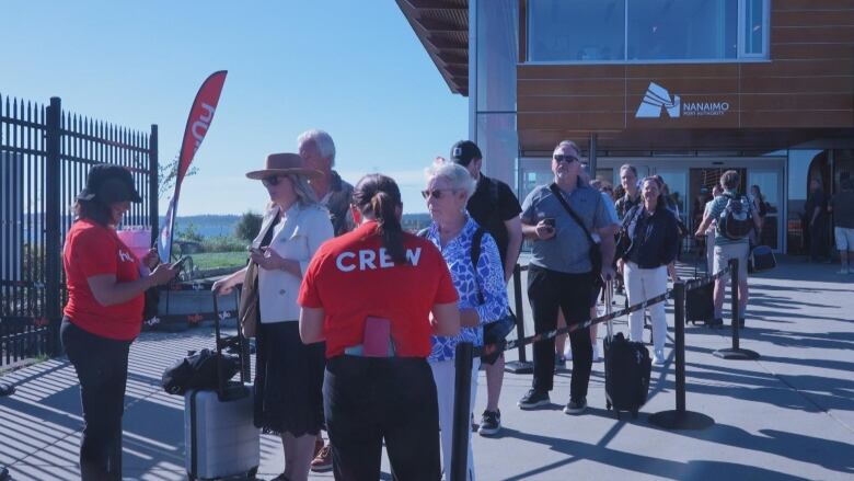 Passengers in a line by the sea with a building that says Nanaimo in the background.