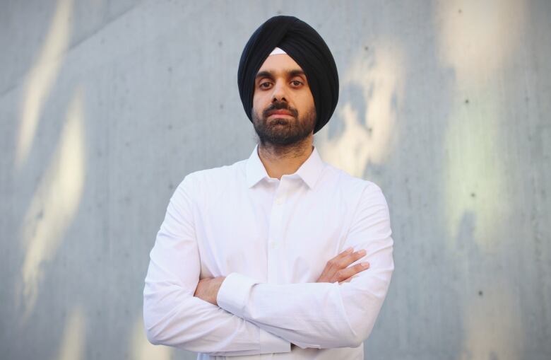 A man wearing a black turban and white shirt stands with his arms crossed looking at the camera. 