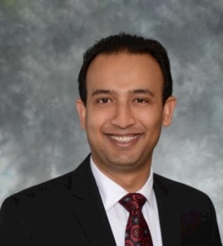 A man in black suit and burgundy tie looks at the camera smiling. 