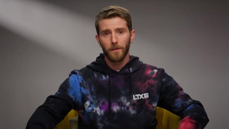 A man with short brown hair and a beard sits on a chair. He's wearing a tie die pattered hoodie.